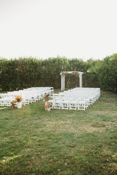 an outdoor ceremony set up with white chairs and flowers on the grass, surrounded by hedges