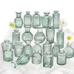 a collection of glass vases and jars on a white cloth with flowers in the background