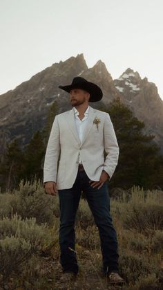 a man in a white jacket and hat standing on a hill with mountains in the background