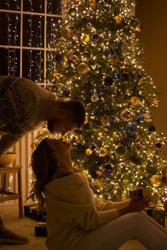 a man and woman kissing in front of a christmas tree with lights all around them