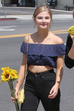 a woman in black pants and crop top holding sunflowers while standing on the street