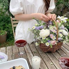 a woman sitting at a table holding a basket filled with food and flowers next to two glasses of wine