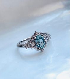 a fancy ring with an aqua and white diamond center surrounded by smaller diamonds on a light blue background