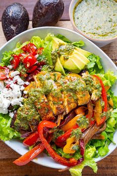 a white bowl filled with vegetables and meat on top of a wooden table next to avocado