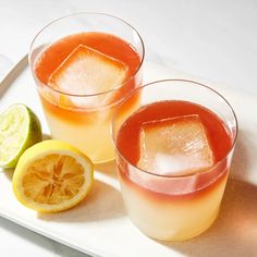 two glasses filled with drinks sitting on top of a white tray next to sliced lemons