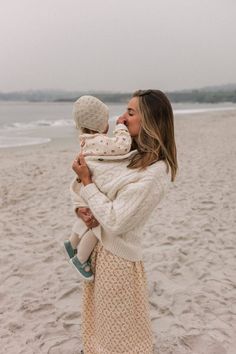 a woman holding a baby in her arms while standing on top of a sandy beach