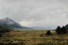 a field with mountains in the background and trees on the other side, under a cloudy sky