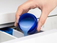 a person pouring blue liquid into a white drawer with drawers in the back ground and one hand reaching for it