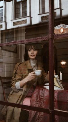 a woman sitting at a table in front of a window drinking from a coffee cup