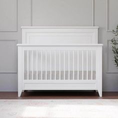 a white crib next to a small plant in front of a gray wall with wood paneling