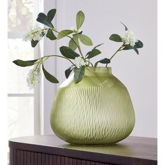 a green vase filled with white flowers on top of a wooden table next to a window