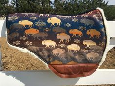 a saddle cover with bison on it is hanging from a white fence in front of some trees