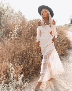 a woman in a white dress and hat standing on a dirt road near tall grass