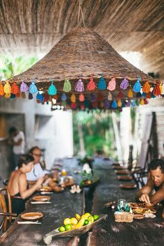 people sitting around a table with food and fruit under a straw umbrella over it that has tassels hanging from the ceiling