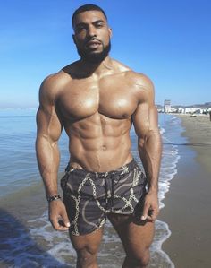 a shirtless man standing on the beach next to the ocean with his hands in his pockets