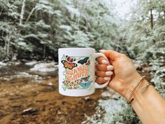 a hand holding a coffee mug in front of a stream and trees with butterflies on it