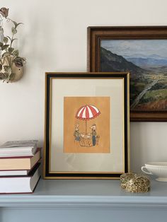 two framed pictures sitting on top of a blue mantle next to books and a vase