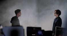 two young men standing next to each other in front of computer monitors on a desk