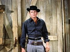 a man wearing a cowboy hat standing in front of a barn door with his hands in his pockets