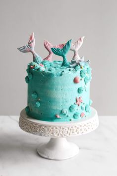 a cake with blue frosting and pink mermaid tail decorations on top, sitting on a white pedestal