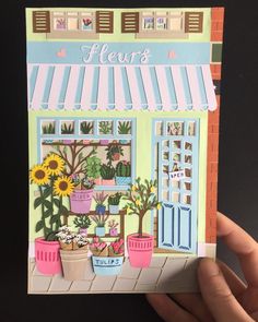 a hand holding up a card with flowers in front of a storefront and potted plants outside