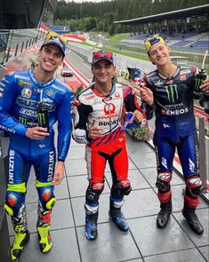 three men standing next to each other on top of a race track with motorcycles behind them