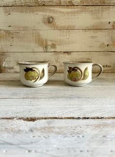 two white cups with designs on them sitting next to each other in front of a wooden wall