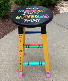 a colorful stool with writing on it sitting in the middle of a sidewalk next to bushes