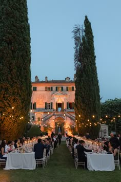 people sitting at tables in front of an old building with tall trees and lights on it