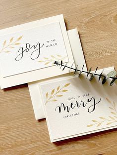 two cards with the words joy to the world and merry christmas written on them sitting on a wooden table