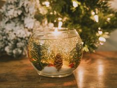 a fish bowl filled with water and pine cones on top of a wooden table next to a christmas tree