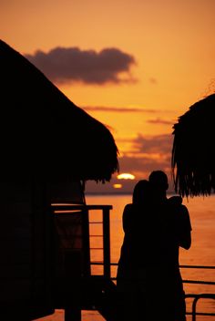 two people standing next to each other in front of the ocean at sunset with thatched umbrellas