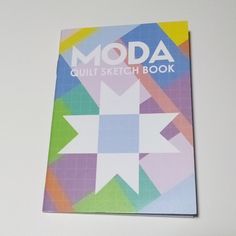 the moda quilt sketch book sits on a white table next to a pen and scissors