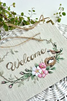 an embroidered welcome sign sitting on top of a wicker basket next to some flowers