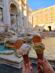 two people holding up ice cream cones in front of a fountain with statues on it