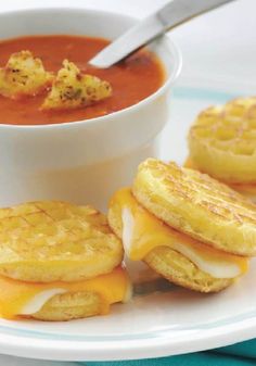 three waffle sandwiches on a plate with a bowl of tomato soup