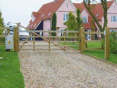 a wooden gate in front of a pink house on a gravel road with grass and trees