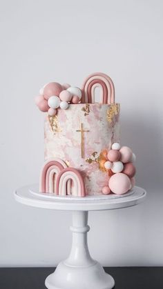 a pink and gold cake with balloons on top is sitting on a white pedestal in front of a gray wall