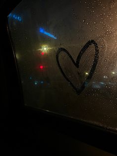 a heart drawn on the side of a window with raindrops and street lights in the background