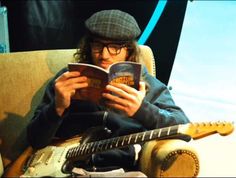 a man sitting in a chair reading a book while holding an electric guitar next to him