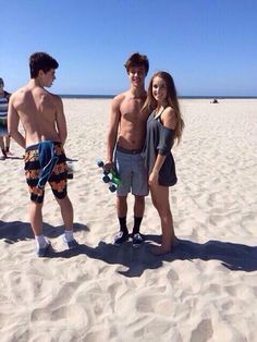 three people standing in the sand with one holding a skateboard and two wearing shorts