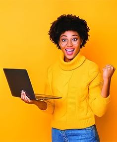 a woman holding a laptop computer in her right hand and smiling at the camera while standing against a yellow wall