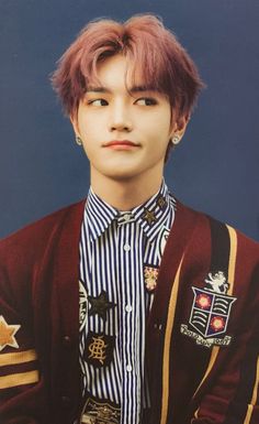 a young man with pink hair wearing a striped shirt and maroon cardigan jacket, standing in front of a blue background