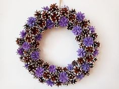 a wreath with pine cones and purple flowers hanging on a wall in front of a white background