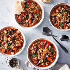 three bowls of pasta soup on a table with bread and spoons next to it