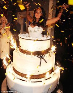 a woman standing in front of a large white cake with sparklers on it and confetti falling from the top