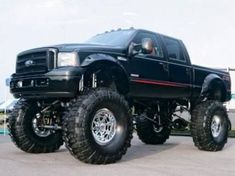 a black truck with big tires parked in a parking lot next to a fenced off area