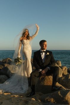 a bride and groom sitting on rocks at the beach with their arms in the air