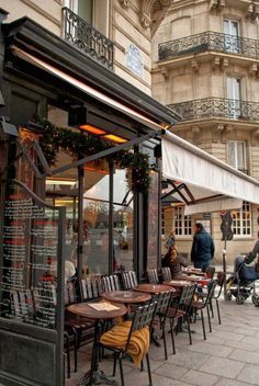 an outdoor cafe with tables and chairs on the sidewalk in front of a tall building