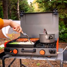 a person cooking food on top of a grill in front of a tent with trees